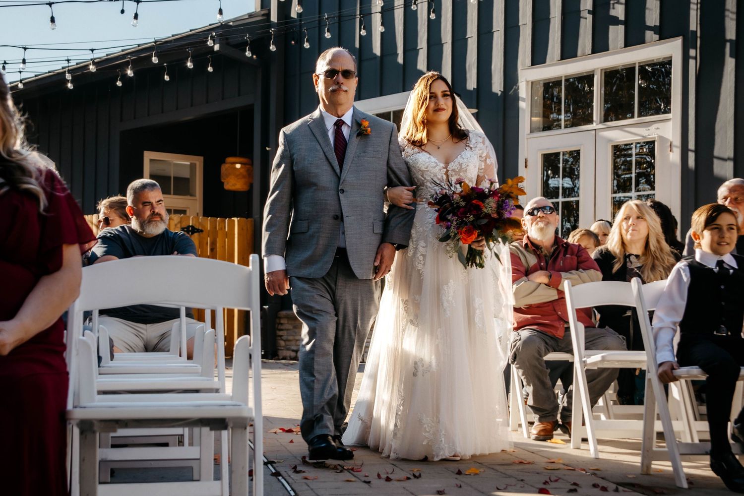 Couple walks down the aisle during outdoor autumn wedding ceremony with string lights and white chairs.