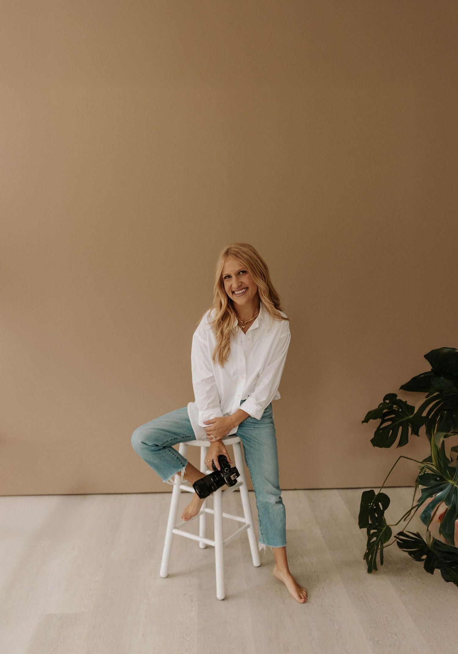 Portrait of a person in a white button-down shirt and light jeans sitting casually on a white stool against a neutral backdrop.