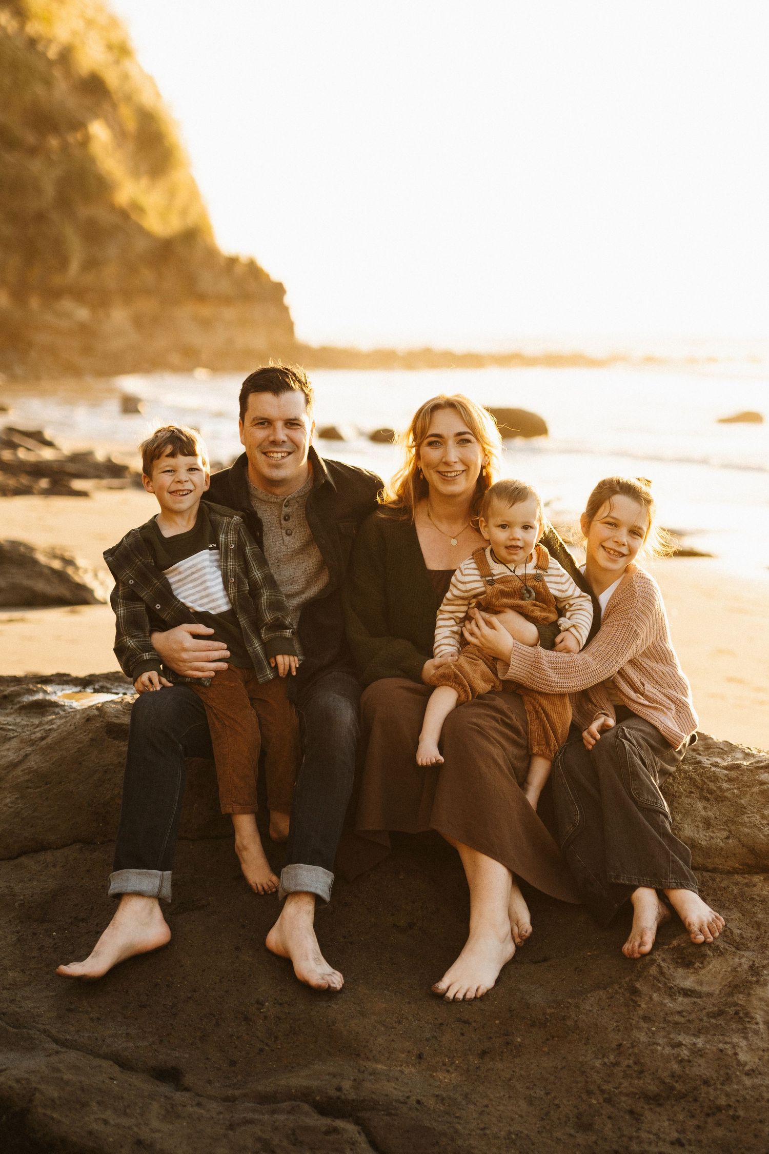 Family of five sits together barefoot on rocky beach during sunset photoshoot.