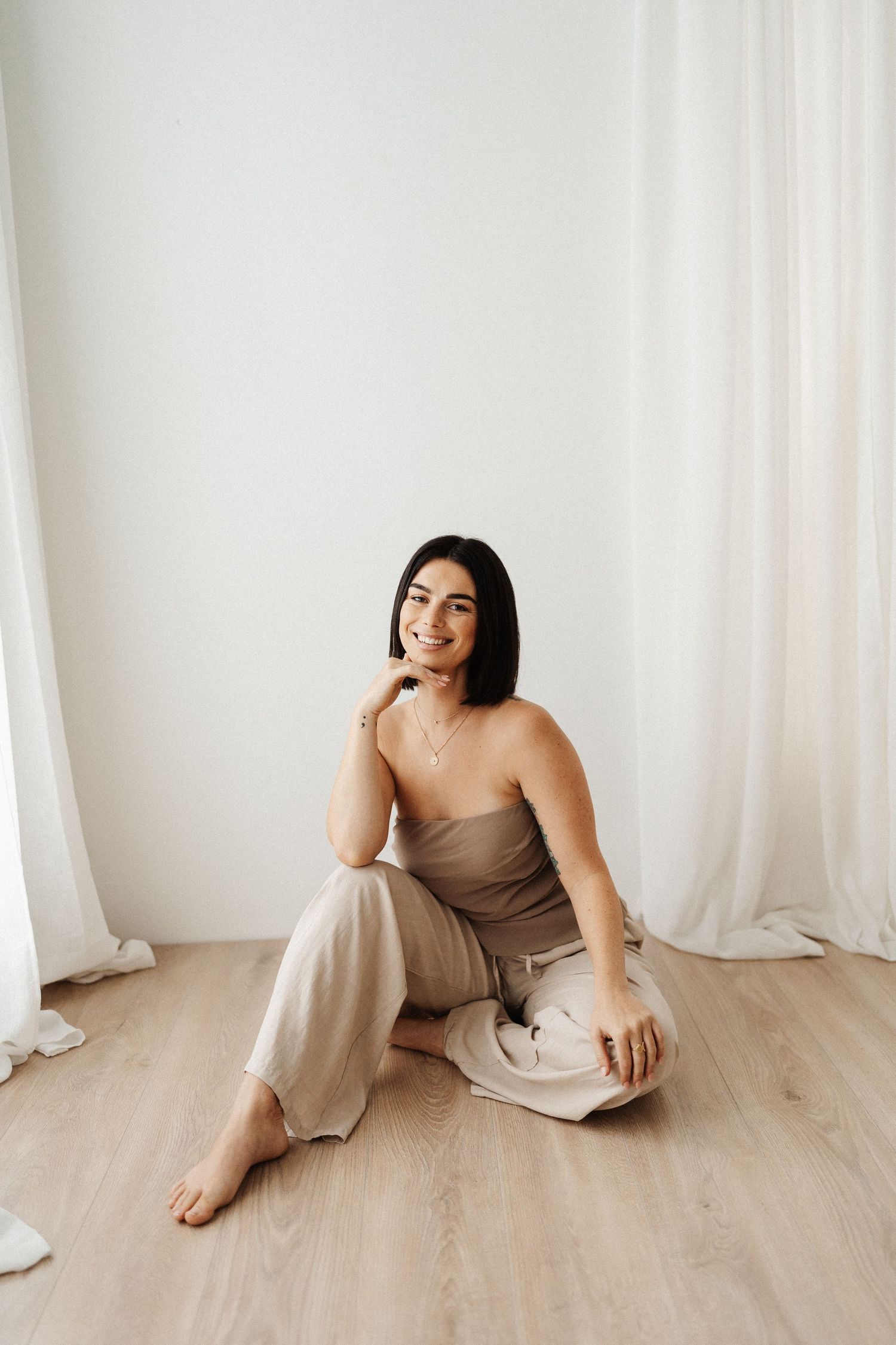 A person in a beige outfit sits on a light wooden floor against a white curtain backdrop in a minimalist setting.