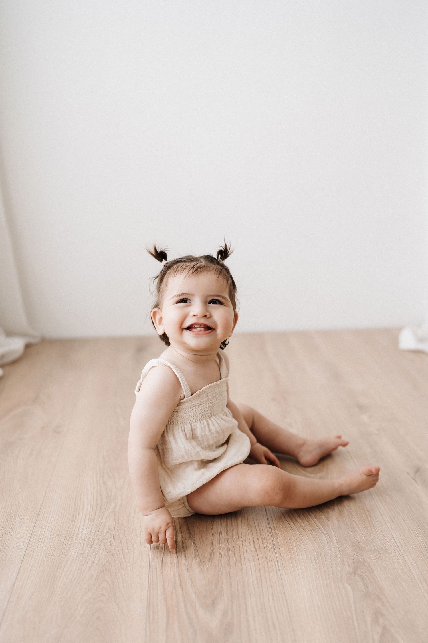 Baby in light colored outfit lying on wooden floor with a joyful smile.