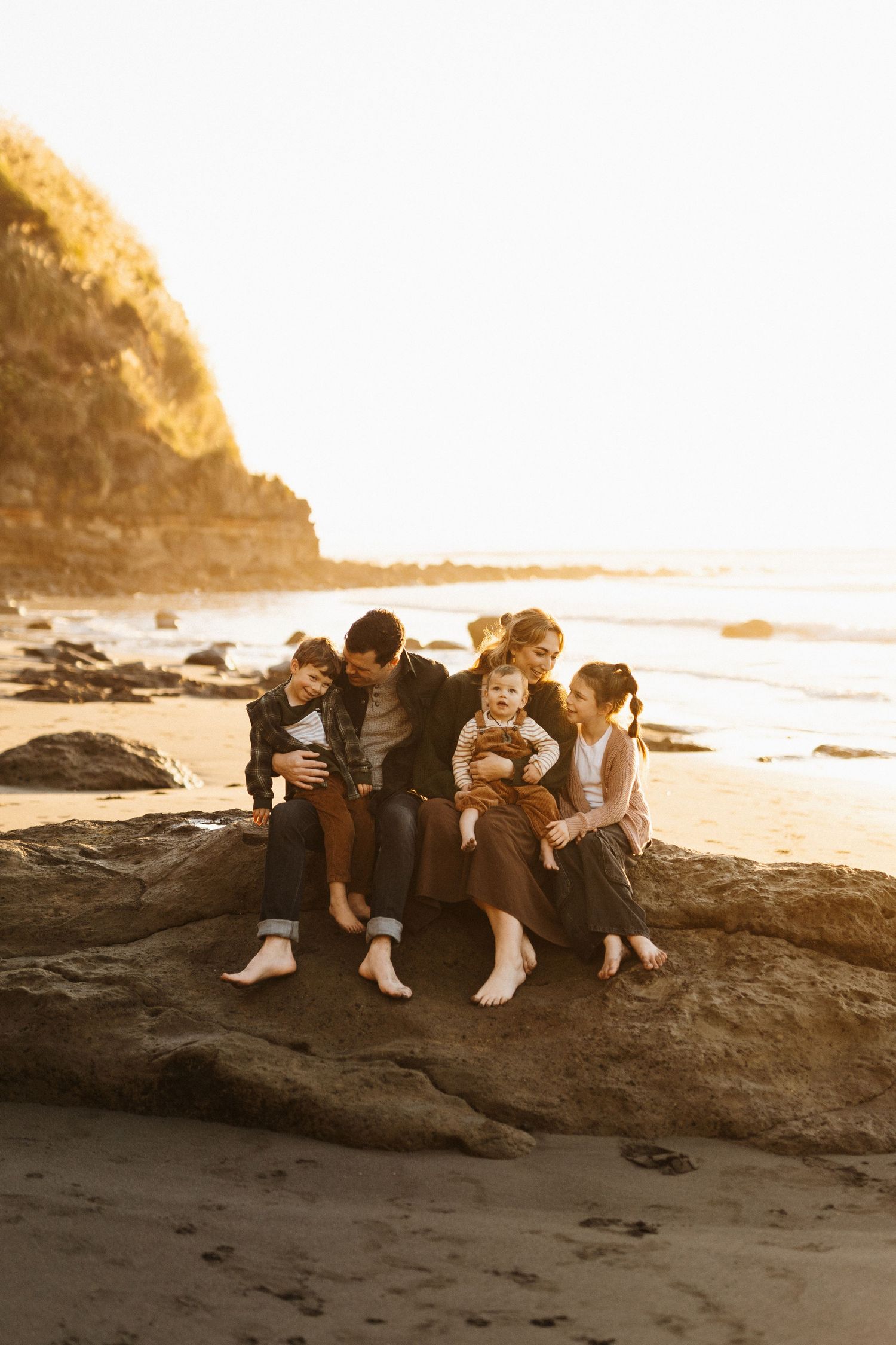 Family group sits close together on coastal rock formation at sunset.