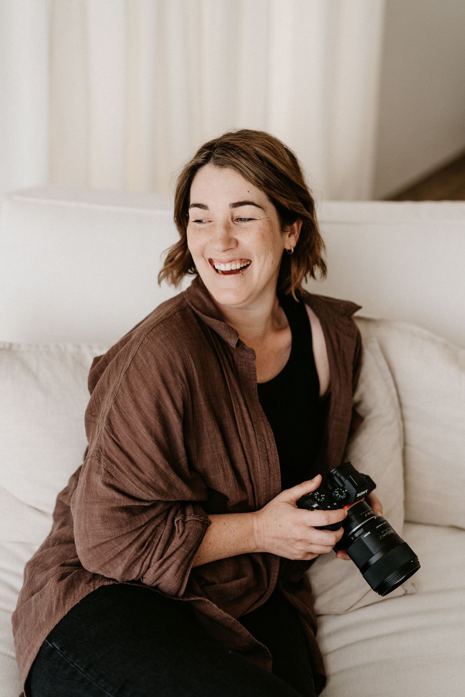 Someone wearing a brown robe lies on white bedding while holding a camera and laughing joyfully.