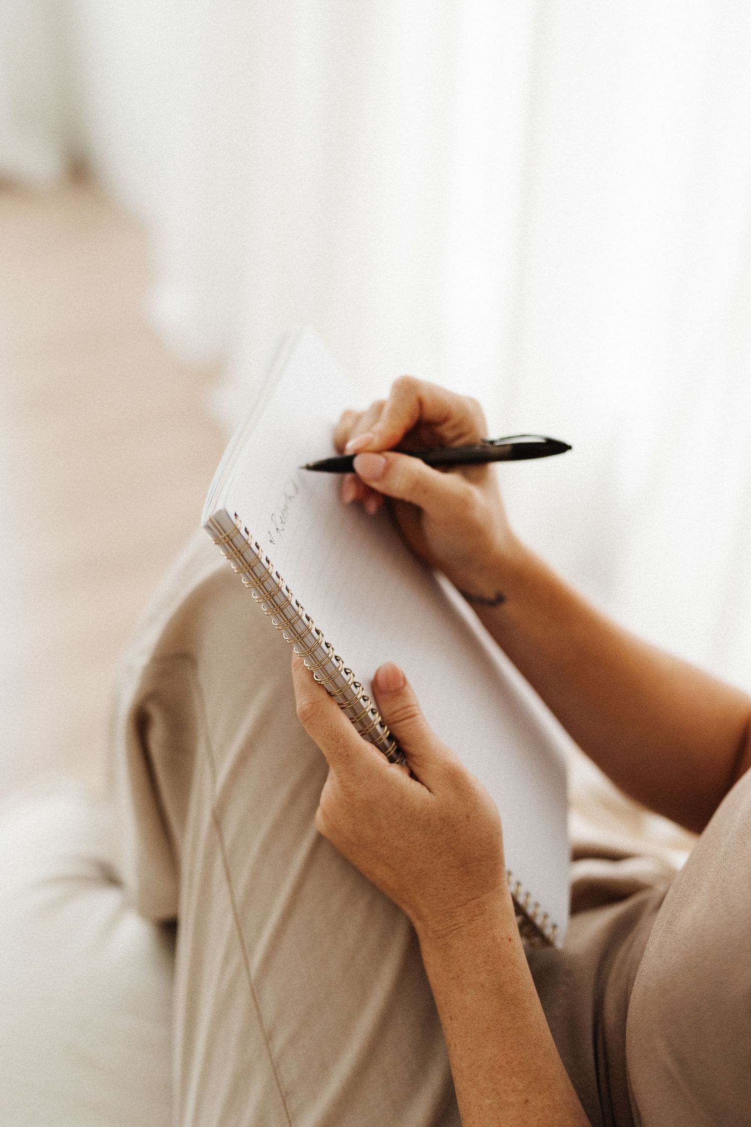 Close-up view of hands writing in a spiral notebook on a white couch, with soft natural lighting in the background.