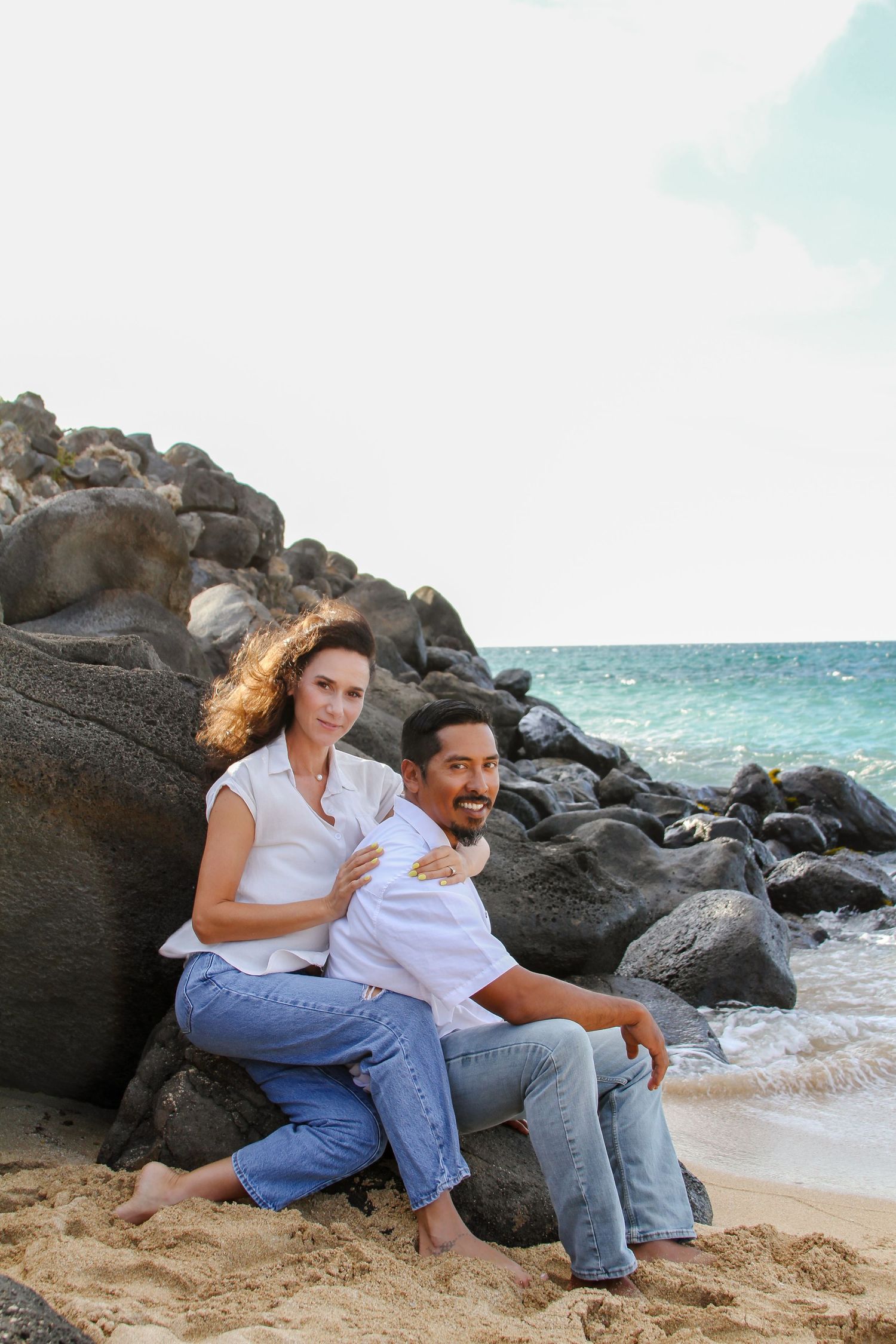A couple sitting together on rocky beach with ocean waves and volcanic rocks in the background.