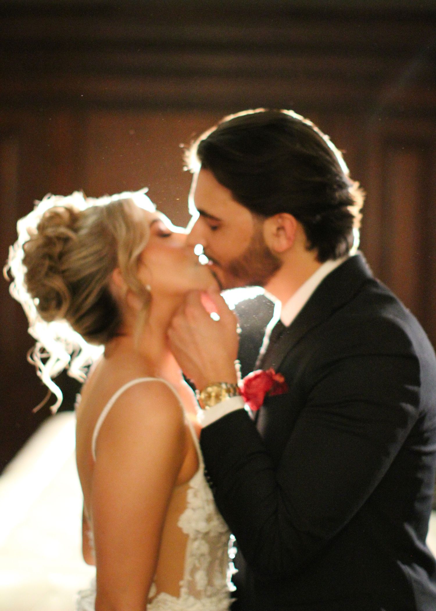 A backlit wedding couple shares a romantic kiss in formal attire.