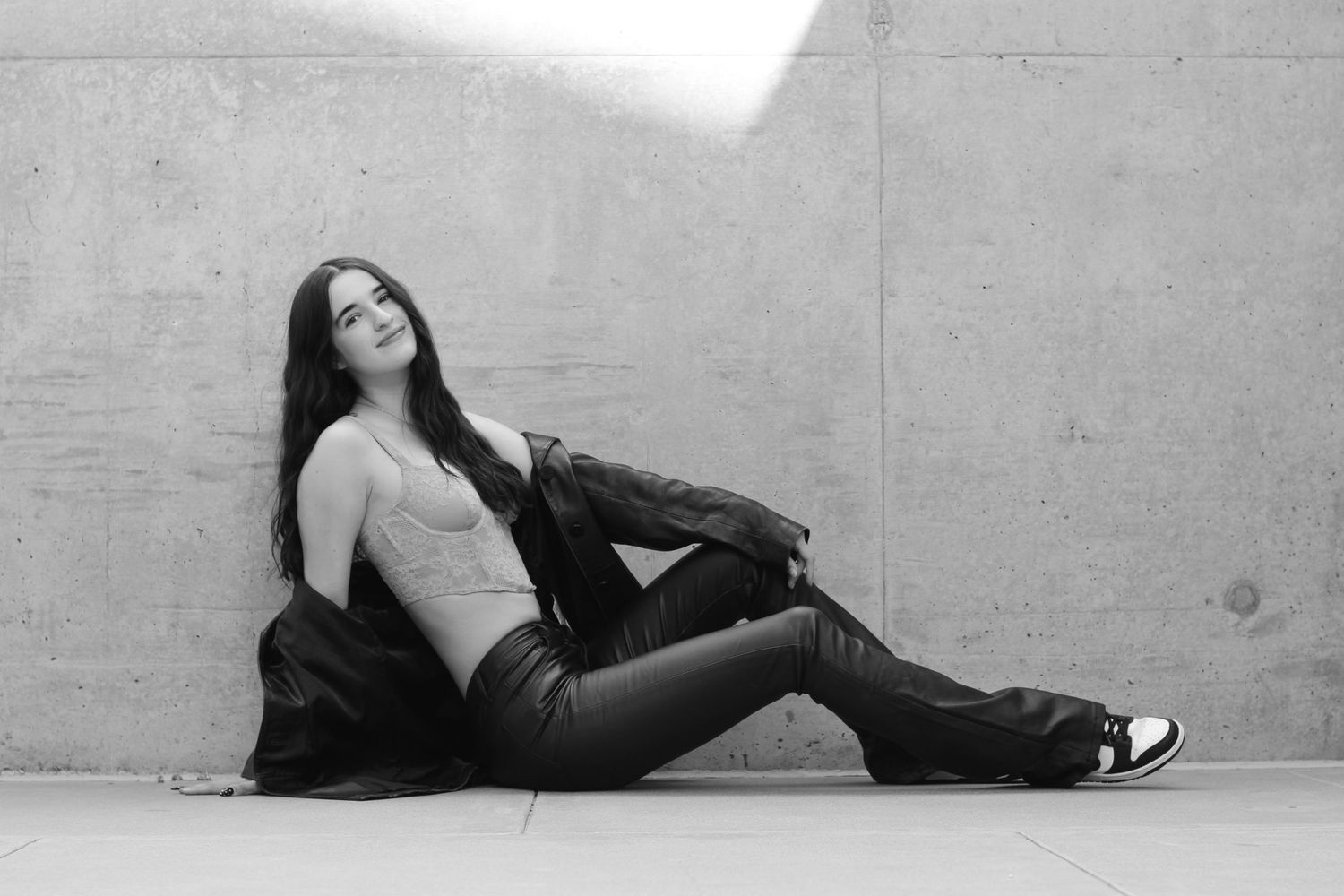 A black and white portrait of someone lounging against a concrete wall in casual attire.