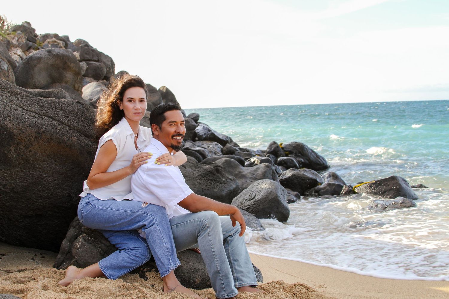 Two people sitting on volcanic rocks at a tropical beach with crashing waves and blue ocean in Hawaii.