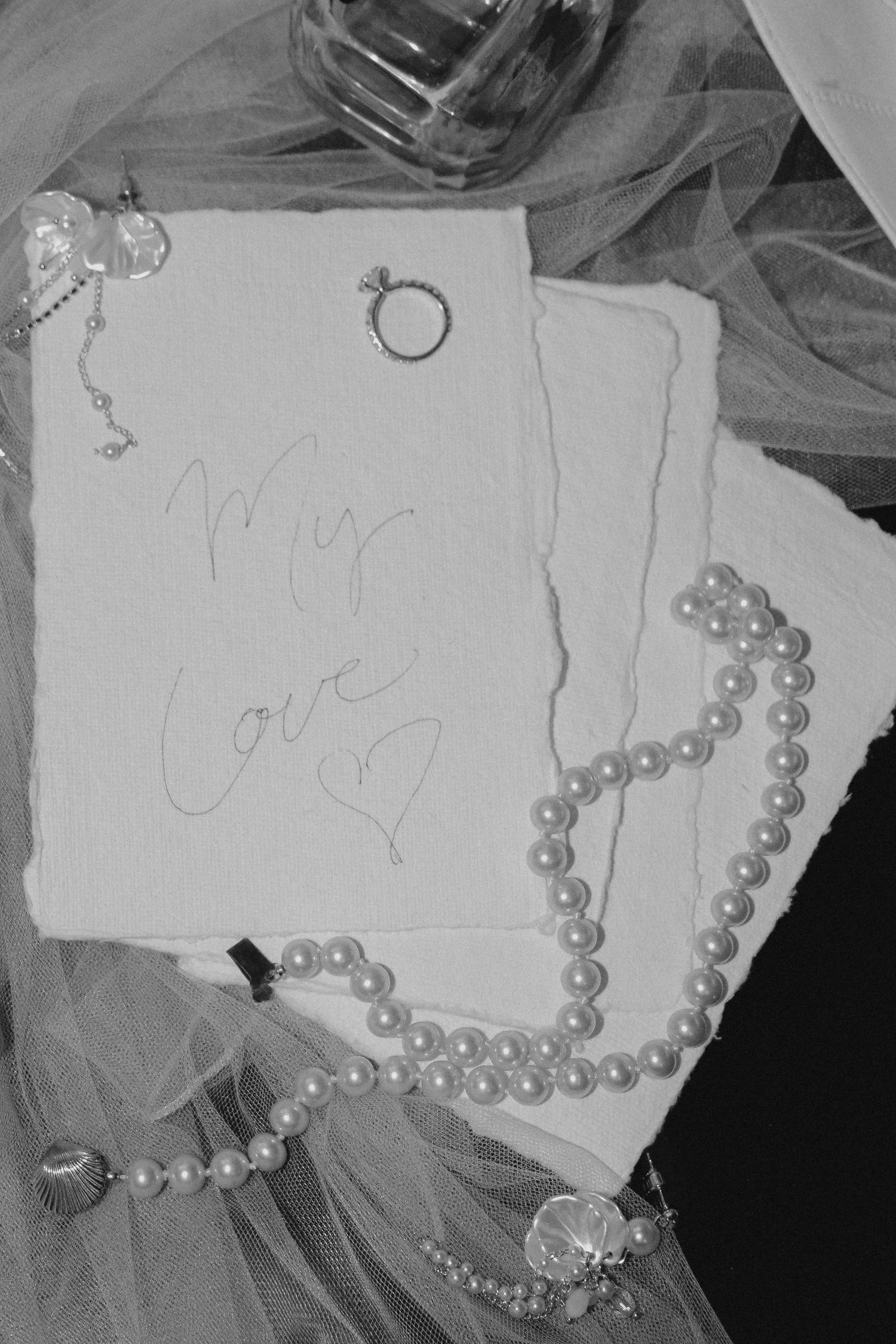 Wedding accessories including pearls, ring, and a handwritten note on white tulle fabric.