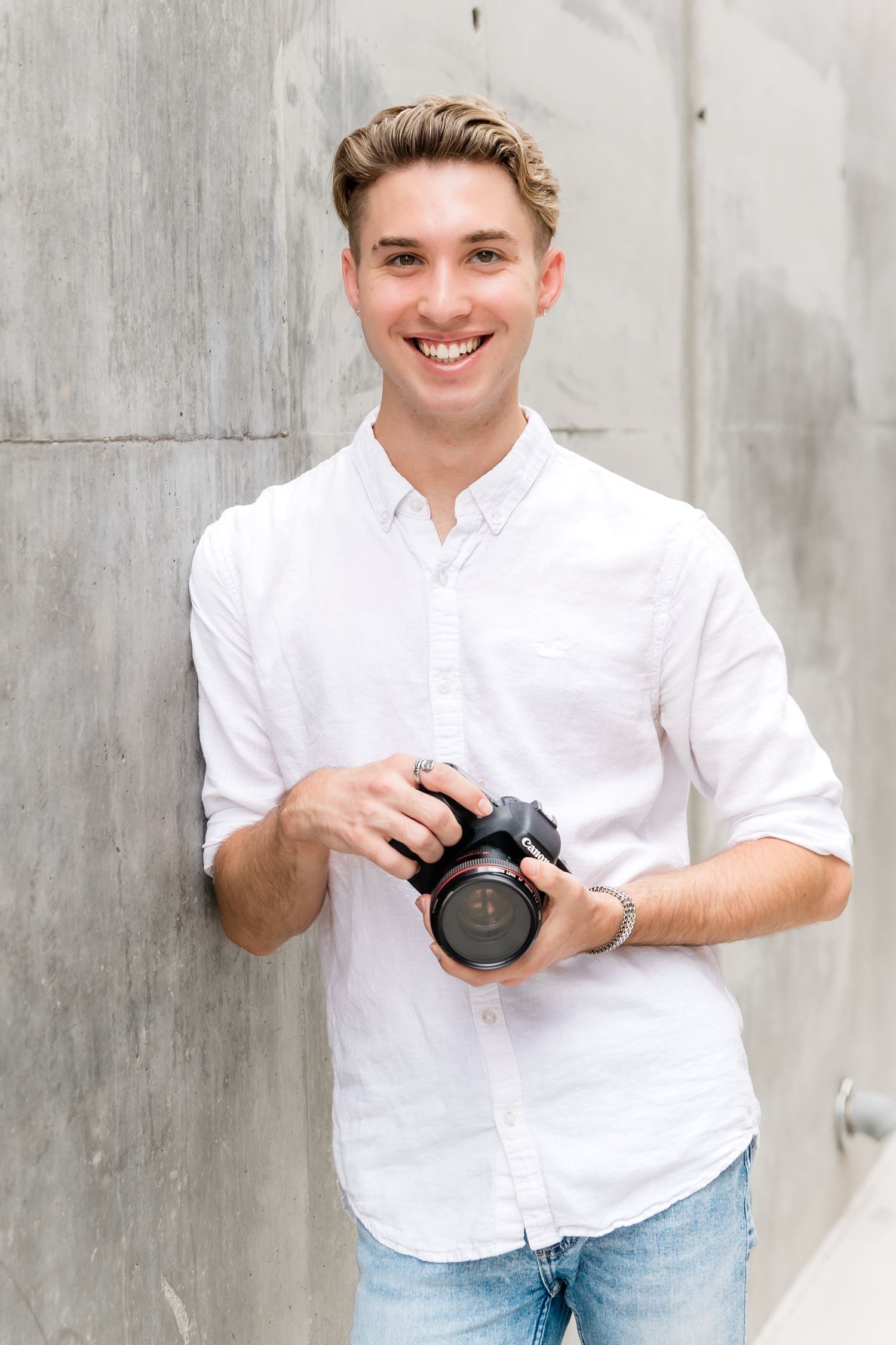 Photographer in crisp white shirt smiling while holding camera against grey wall.