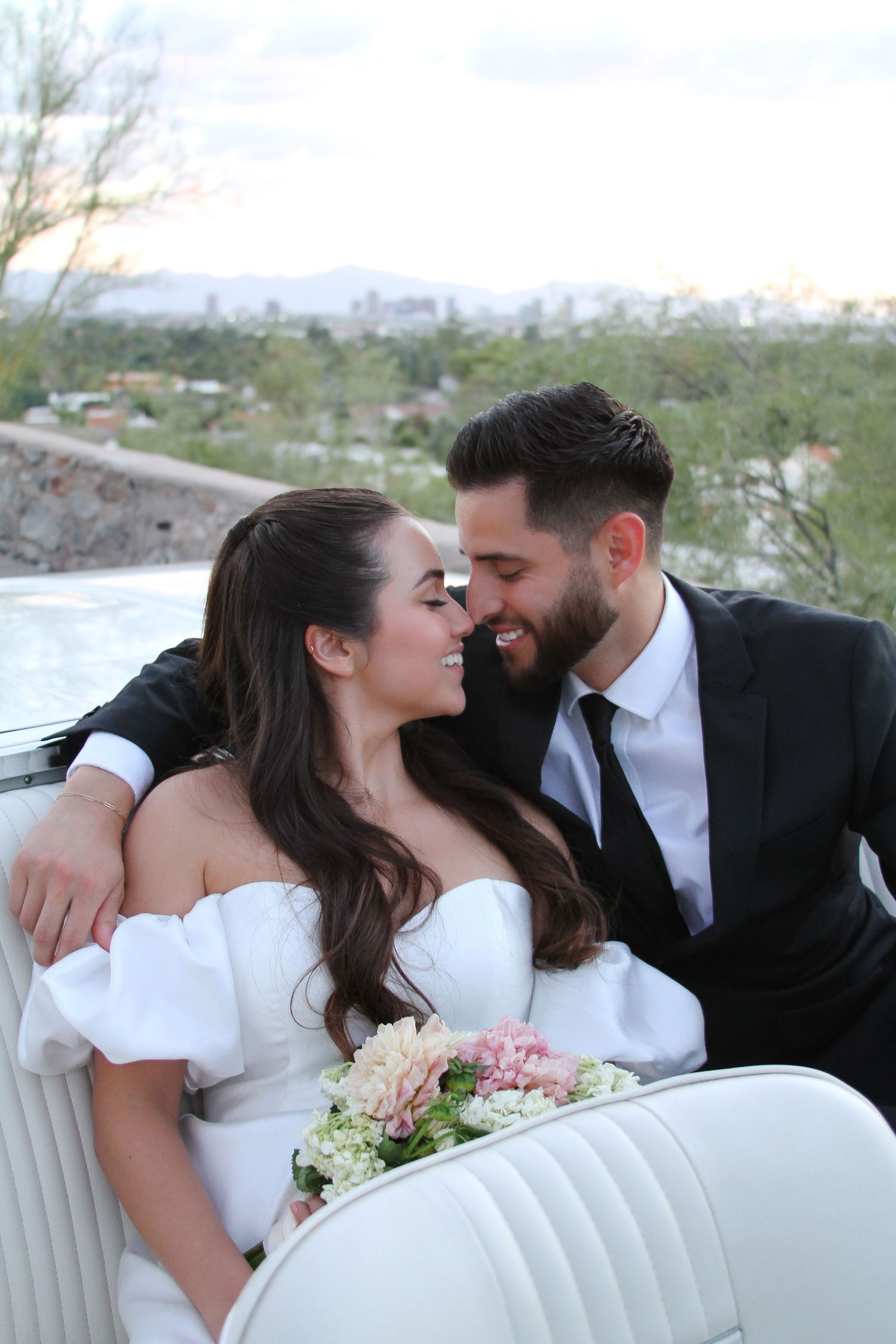 A romantic wedding couple shares an intimate moment in a vintage white car with a bouquet of flowers.
