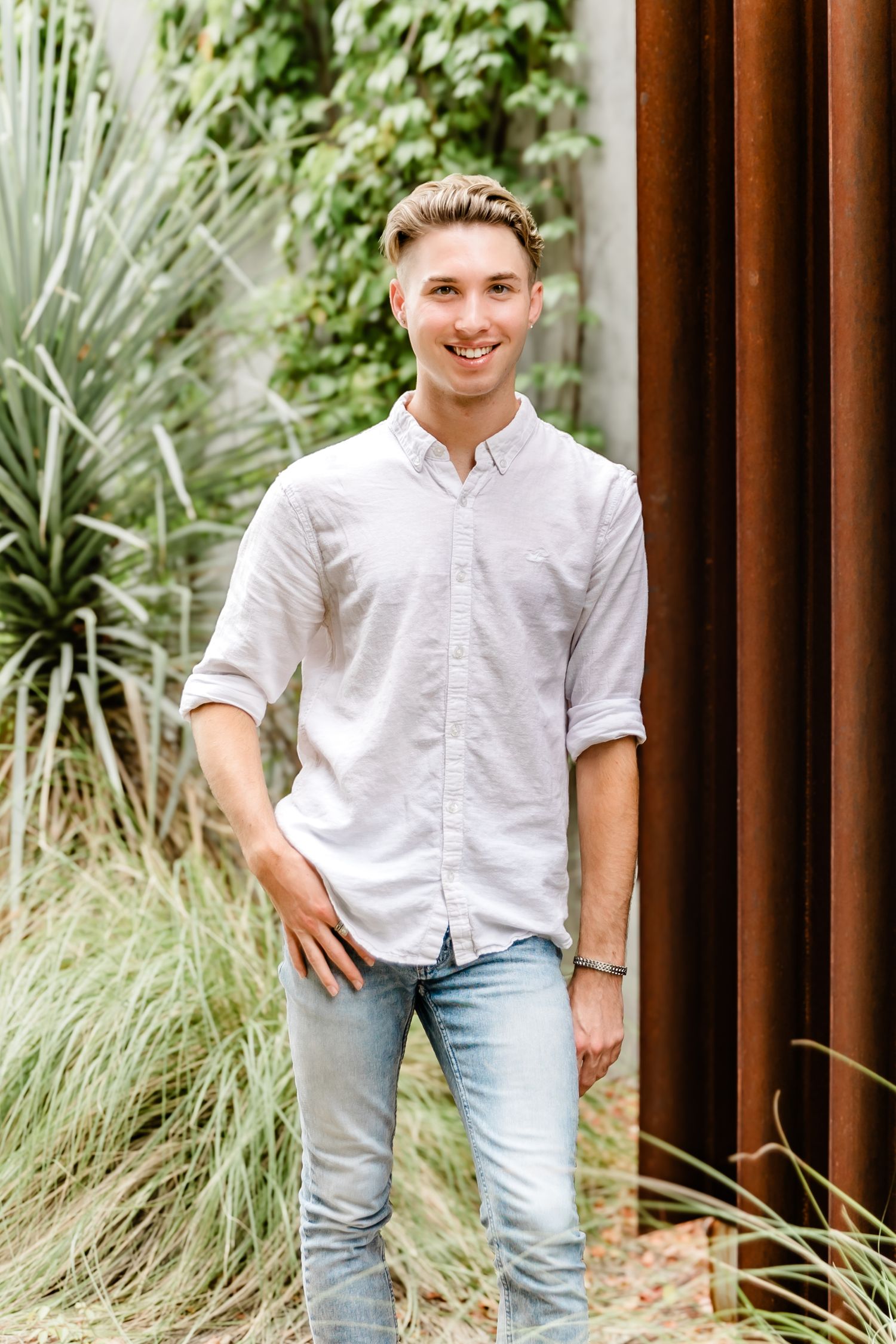 A casual portrait of a young man in a white button-down shirt against a rustic backdrop.