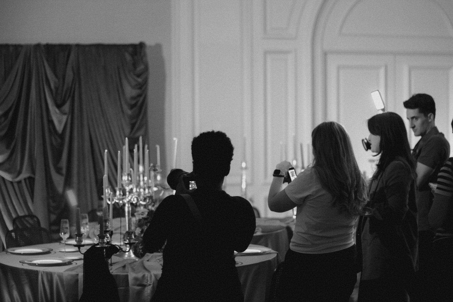 Silhouettes of people gathered in an elegant room with chandeliers and candlelight.