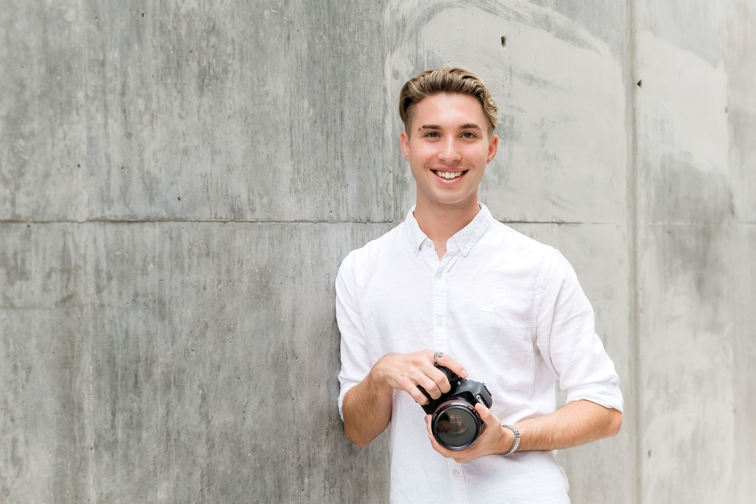Photographer in white button-down shirt poses with camera against concrete background.