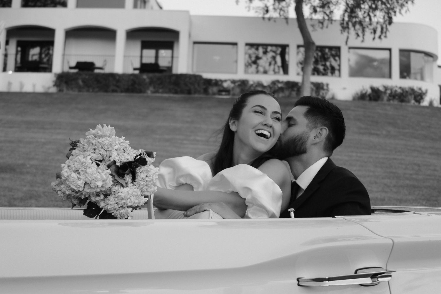A joyful wedding couple laughs together in a classic car during their photoshoot.
