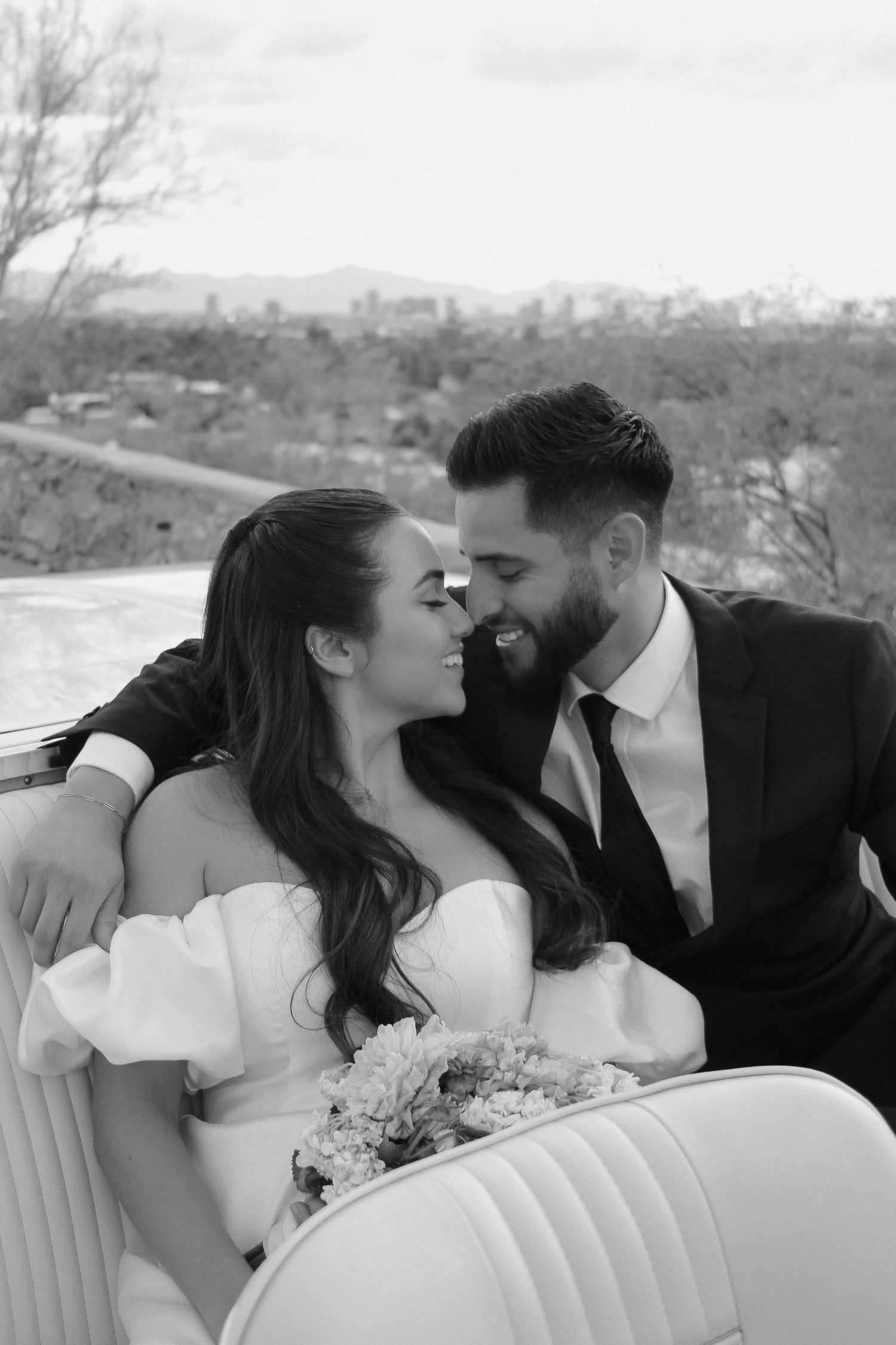 A wedding couple shares an intimate moment in a vintage car with city views.