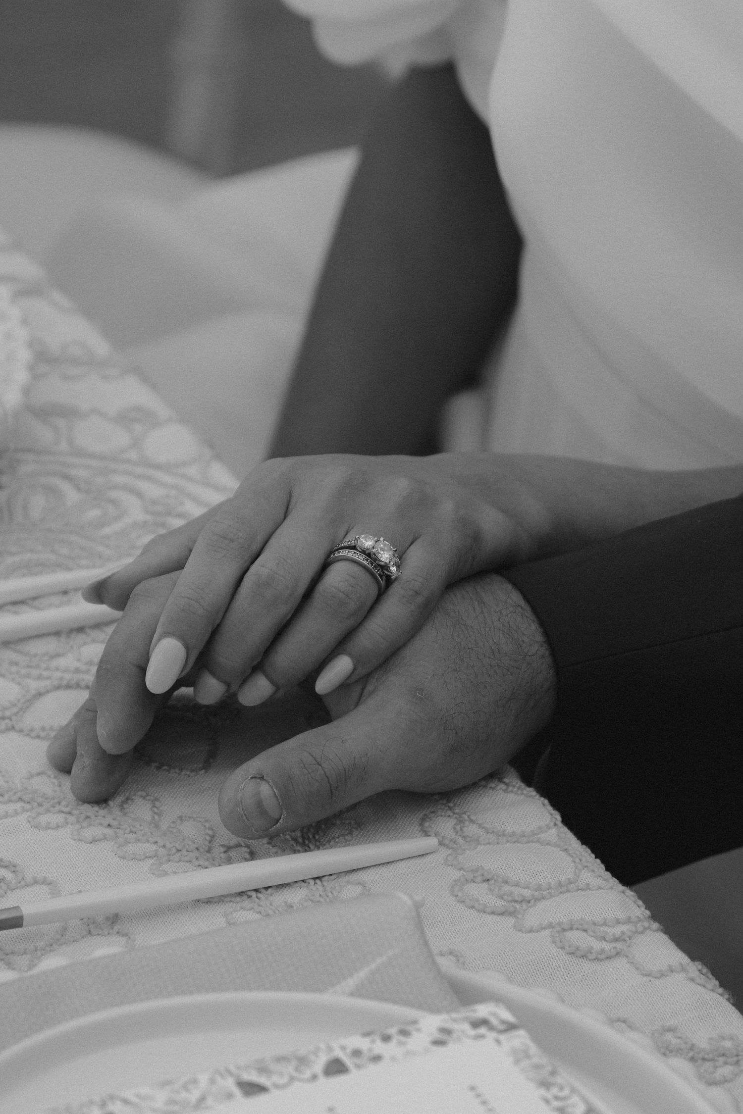 Black and white close-up of hands with wedding rings resting on a lace tablecloth.