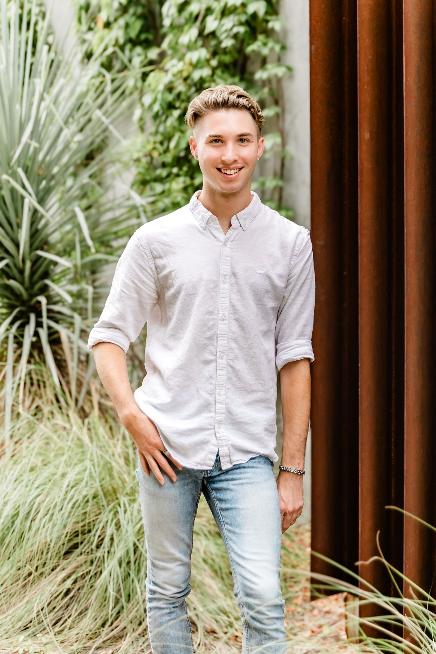 Person in white button down shirt and jeans standing by tropical plants and modern wall.