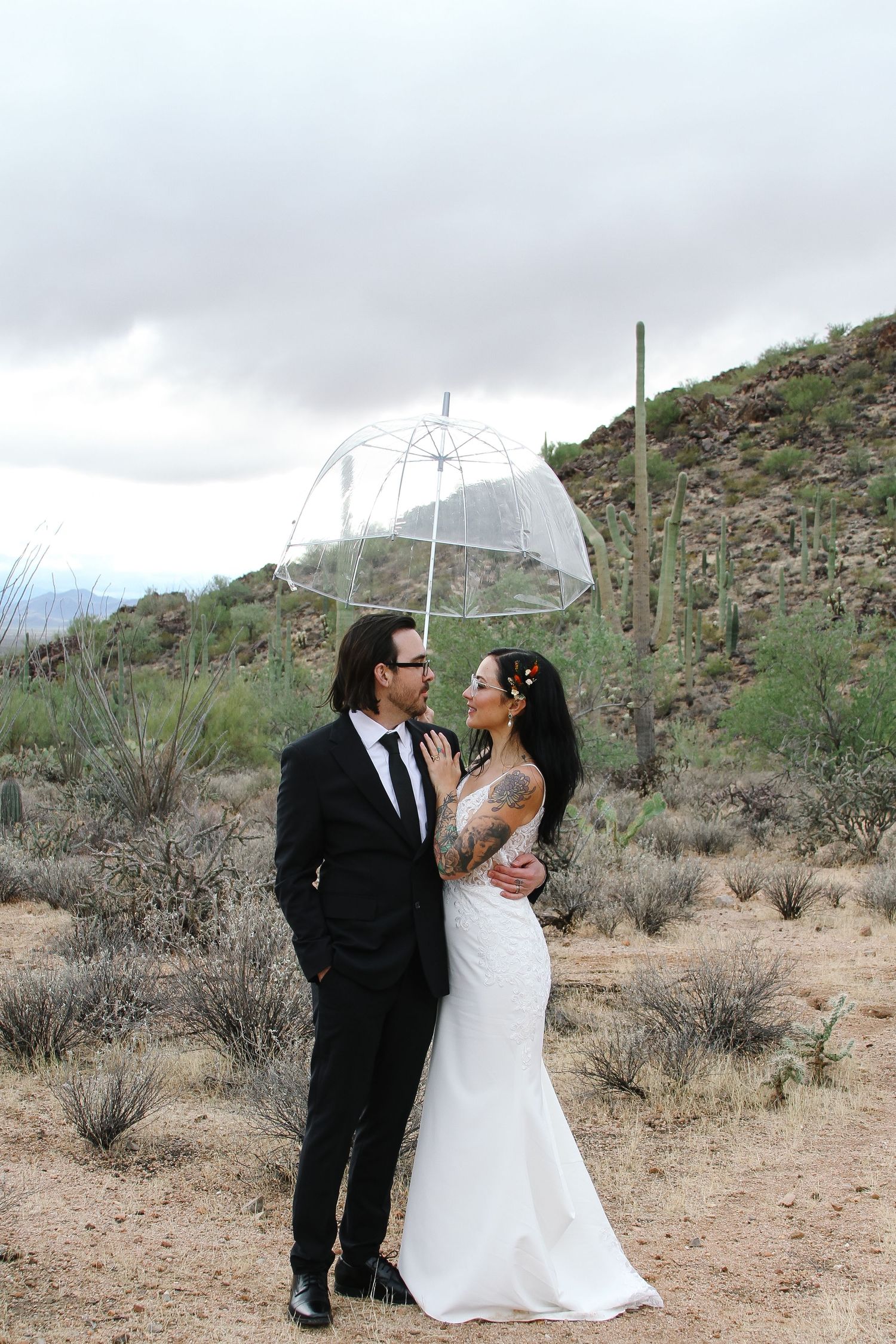 A wedding couple poses with a clear umbrella in a desert landscape with towering cacti.