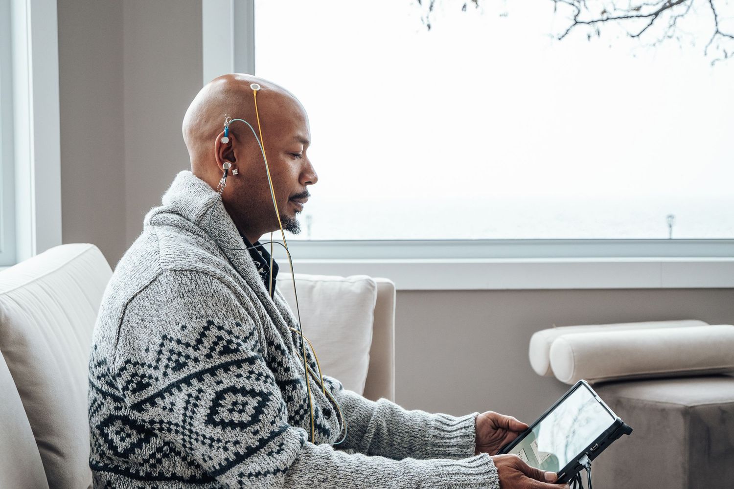 Person wearing grey patterned sweater using tablet device while listening with earbuds near window.