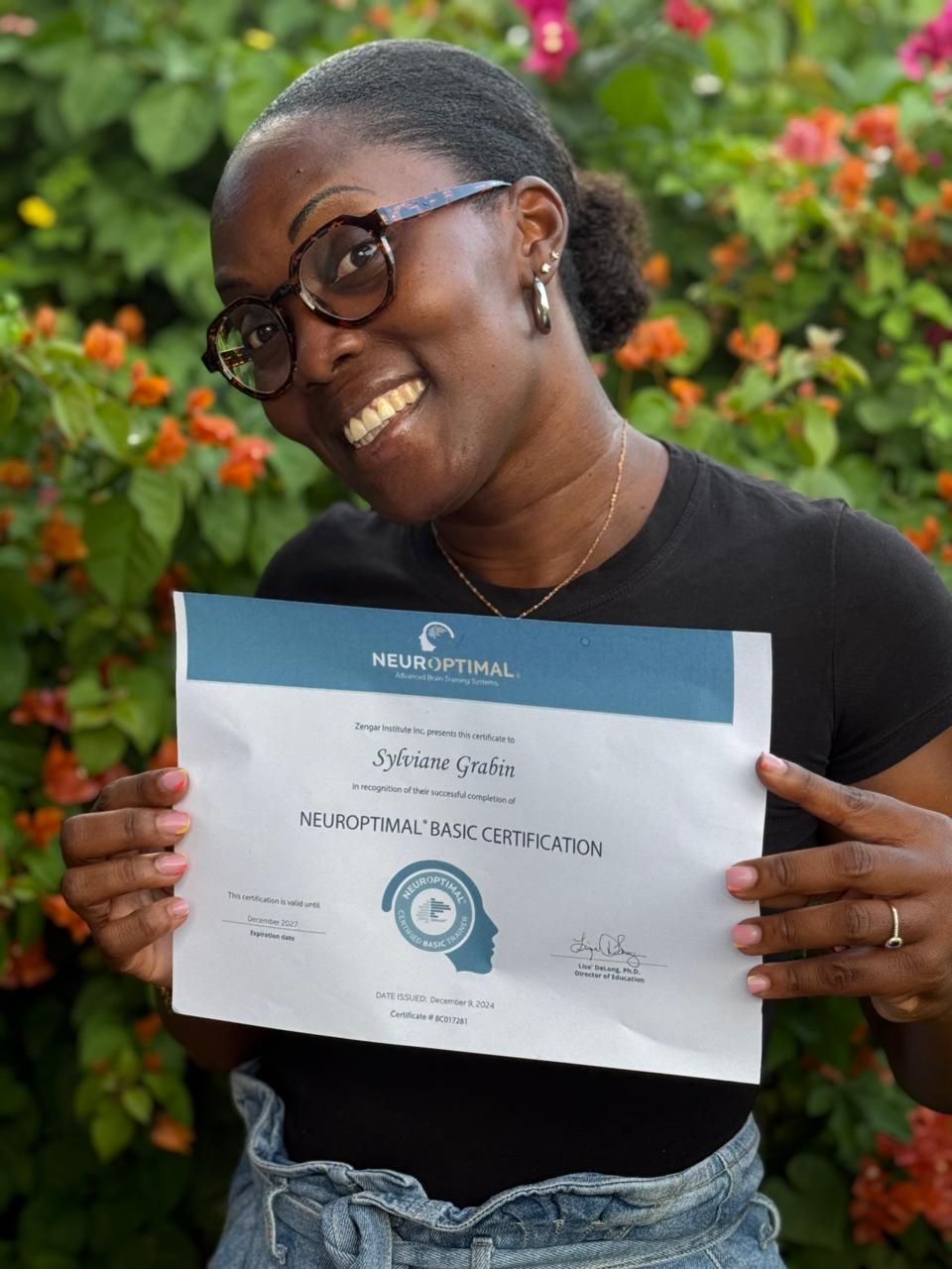 Graduate showing NeuroOptimal certification outdoors with colorful flowers in background.