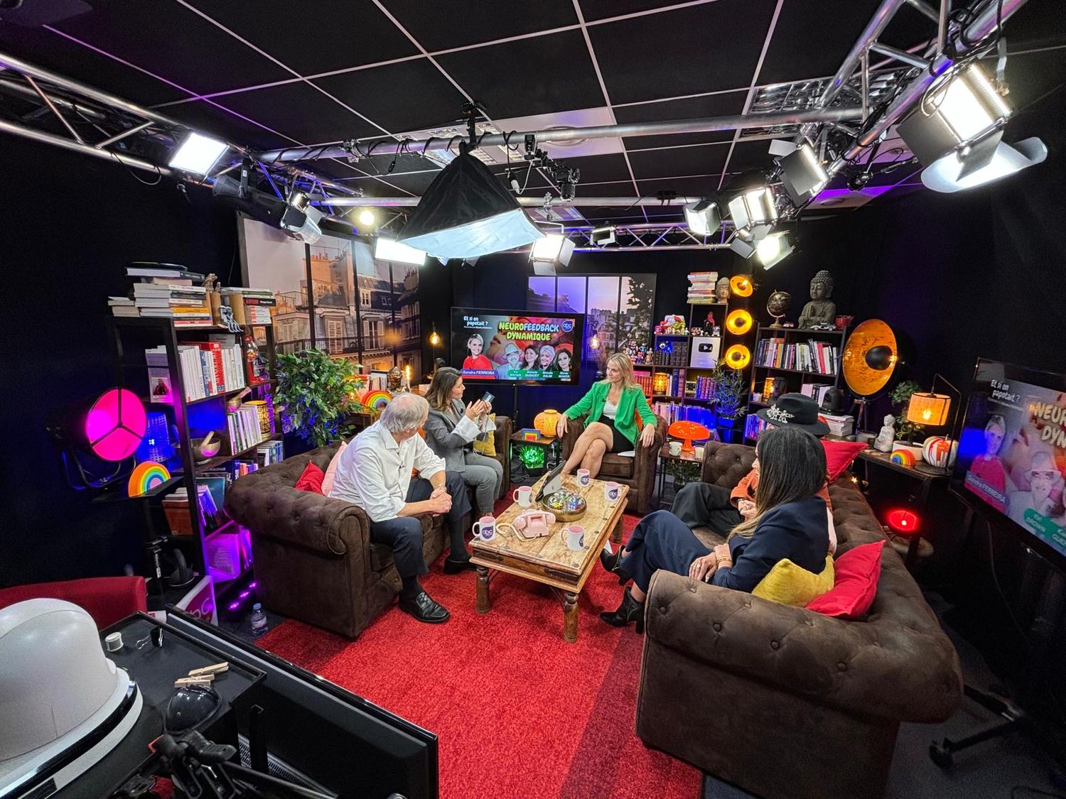 A colorful studio ABC Talk setup with brown couches, neon lights, and gaming equipment arranged on shelves in a black-walled room.
