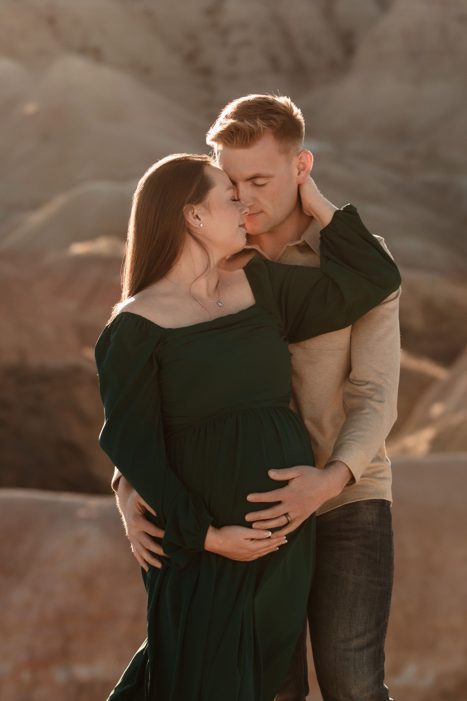 Intimate couple in matching dark green and beige outfits embrace tenderly during sunset in a desert landscape.