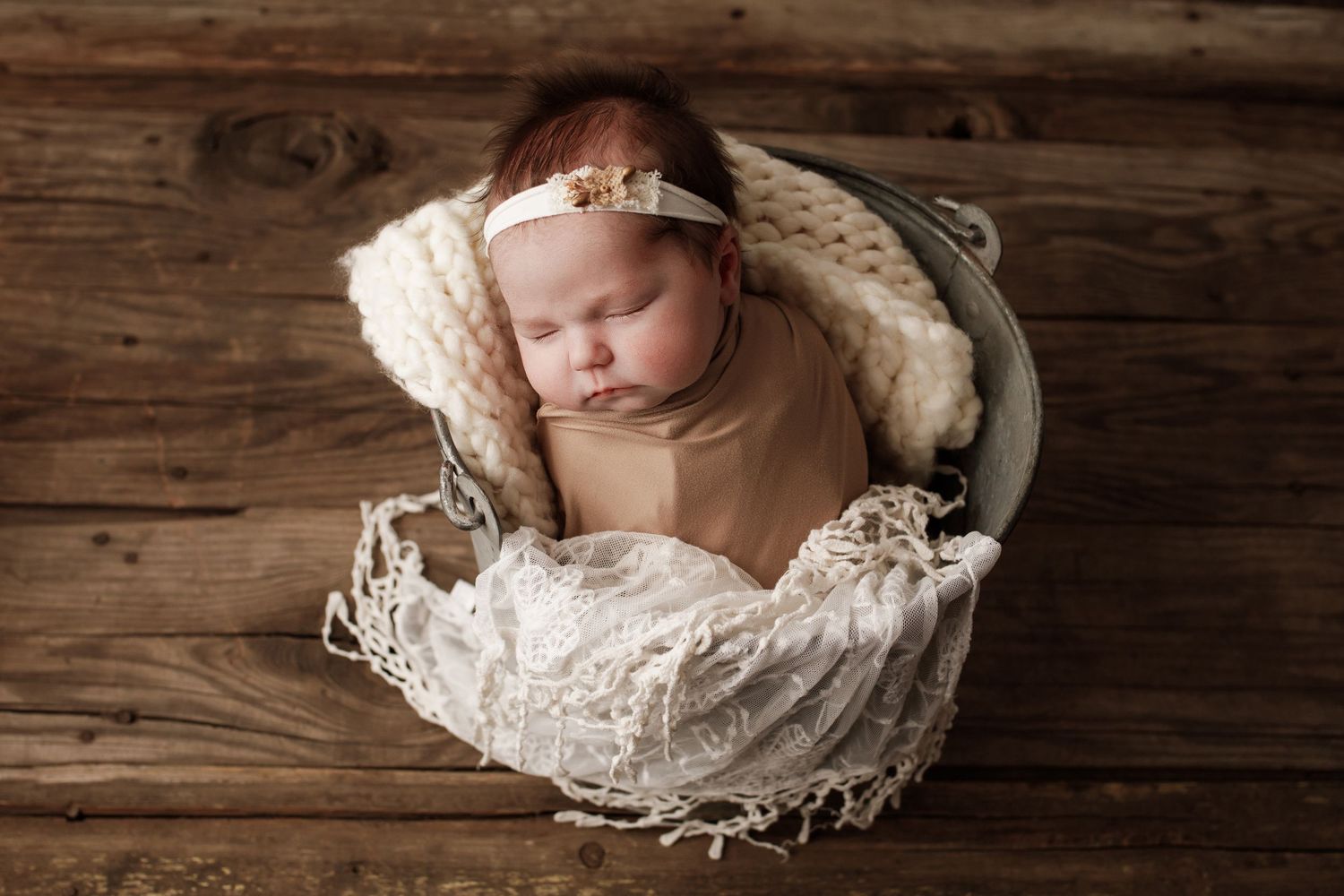 Newborn baby sleeping peacefully in rustic basket with lace and blankets on wooden floor.