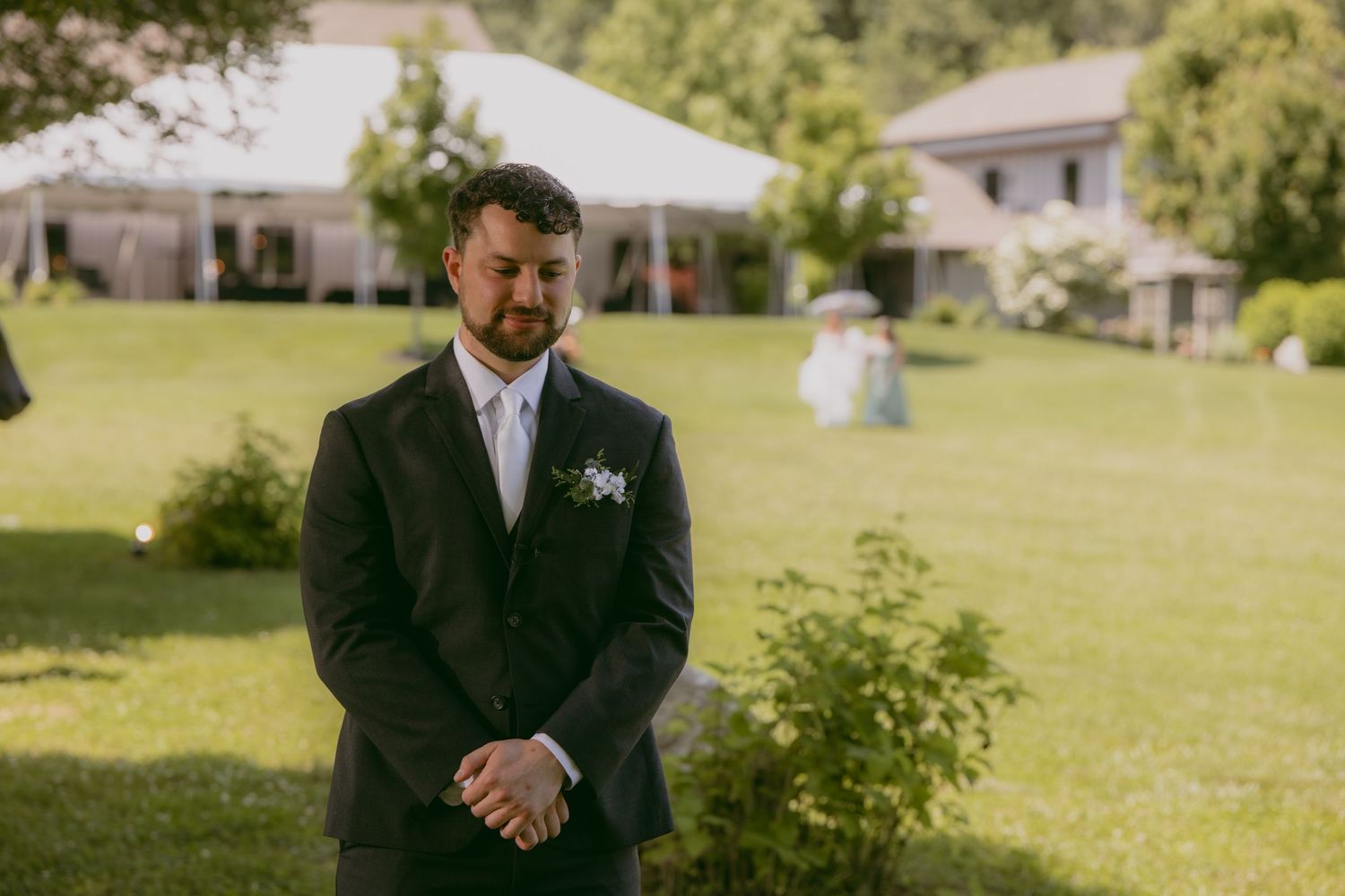 Someone in a dark suit at a garden wedding ceremony with a manicured lawn and white tent in the background.