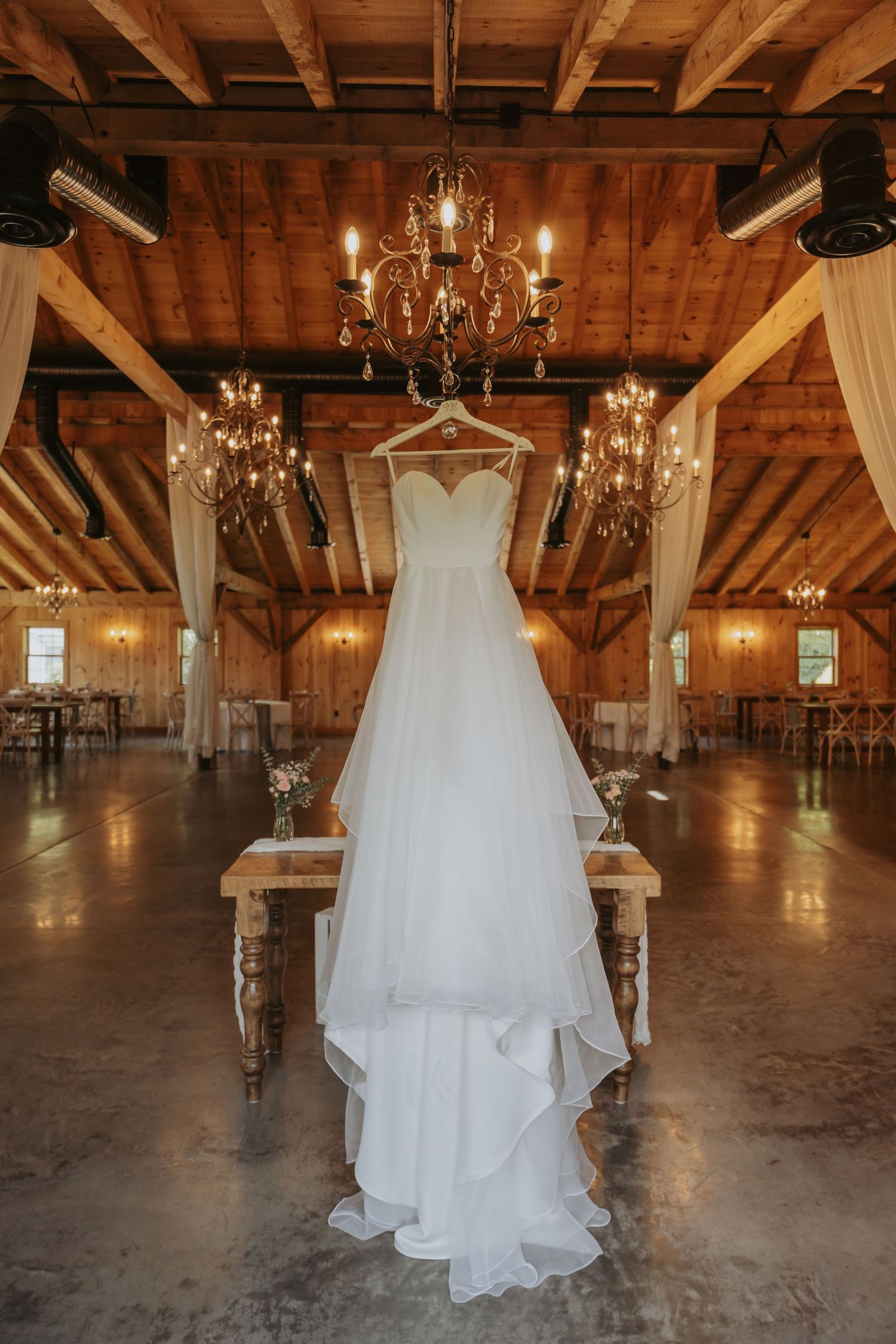 A wedding dress hangs from exposed wooden beams in a rustic barn venue illuminated by elegant crystal chandeliers.