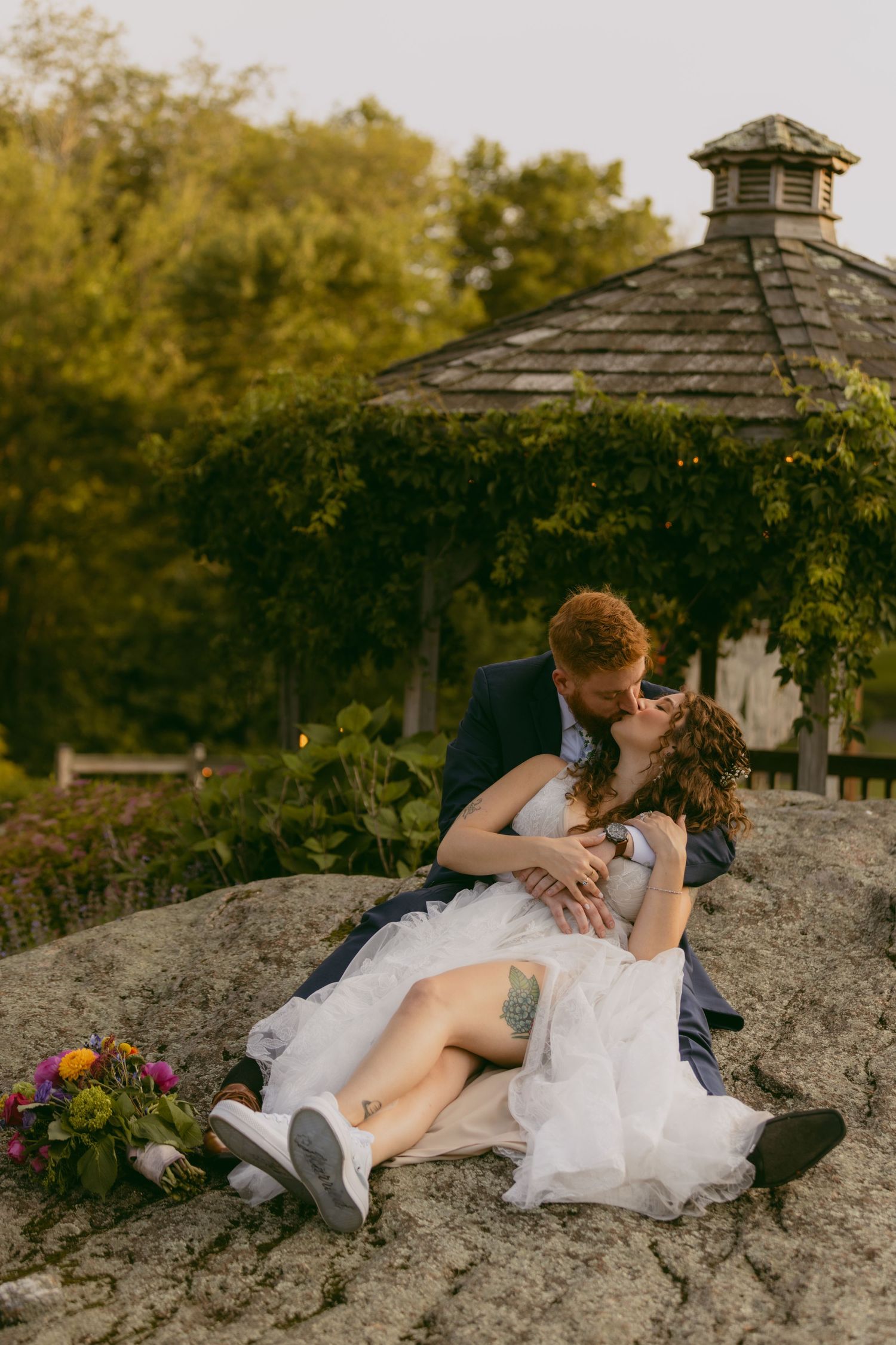 A romantic wedding couple lying together on a rocky outcrop during golden hour with wooden gazebo in background.