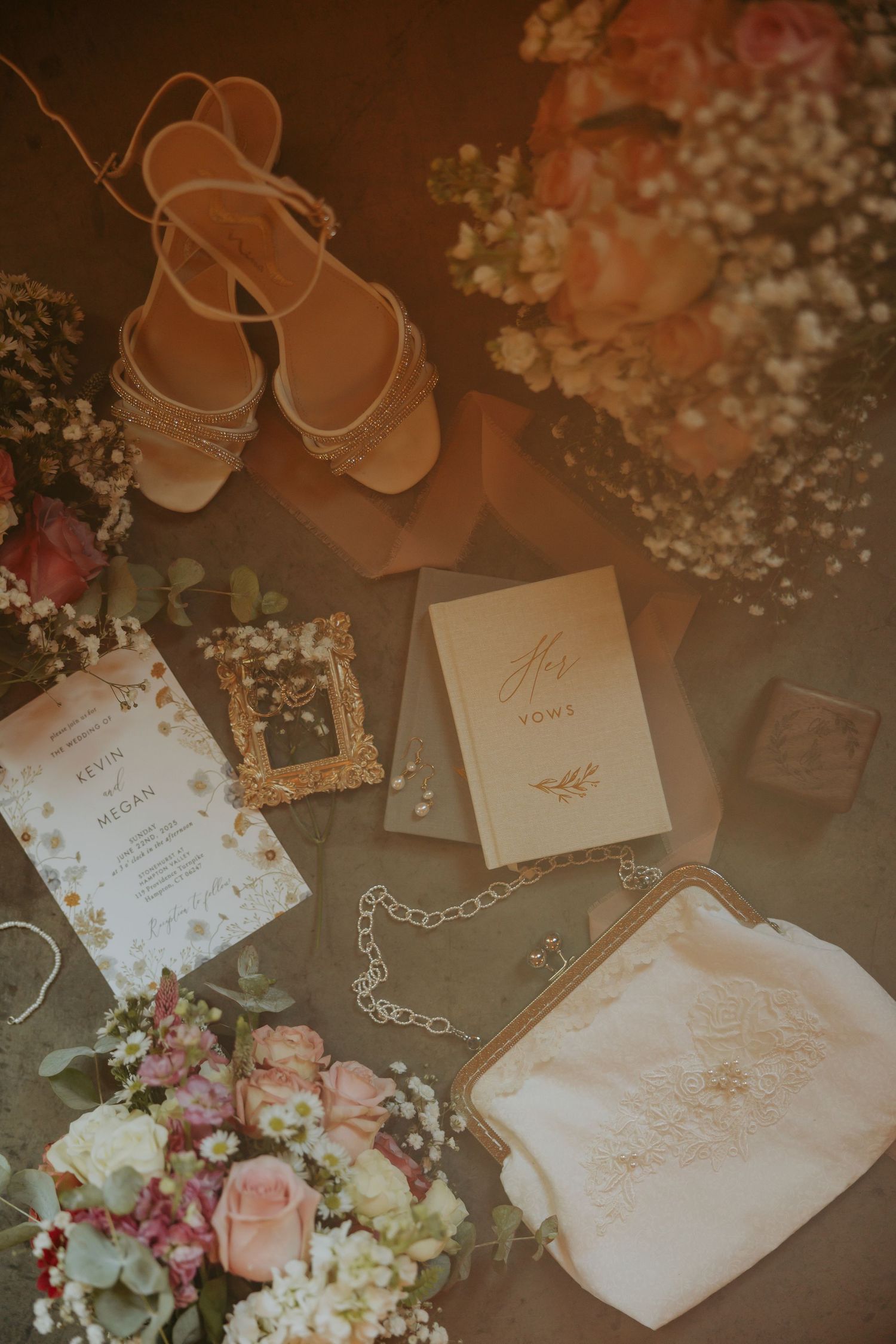 Wedding details flatlay with bridal sandals, invitation cards, jewelry, baby's breath flowers and pink roses in warm lighting.