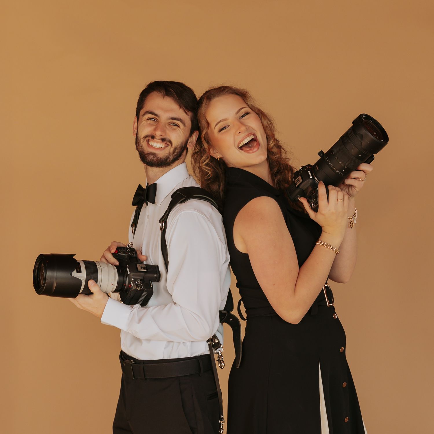 Two photographers in formal attire laugh and pose back-to-back while holding professional cameras with zoom lenses.