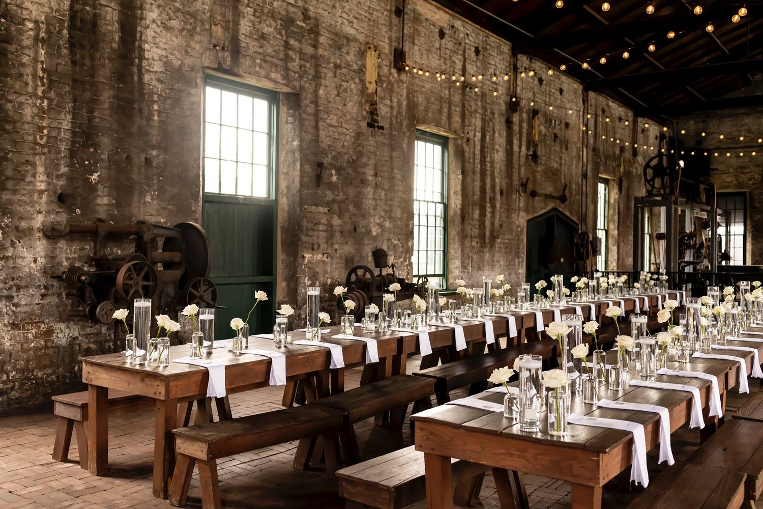 Rustic wedding reception setup in an industrial venue with long wooden tables and string lights hanging overhead.