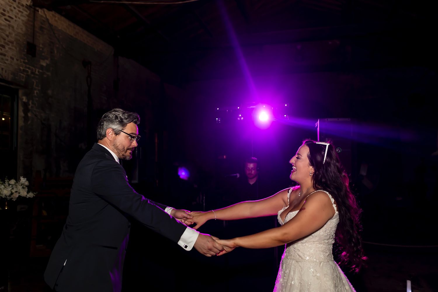 Newlywed couple dances together during their first dance at wedding reception with purple lighting effects in background.