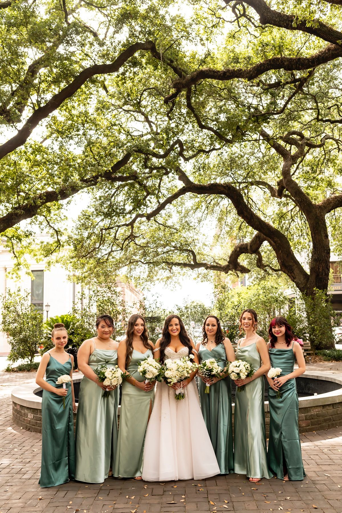 A bridal party stands together in varying shades of sage and mint green dresses under a large oak tree canopy.