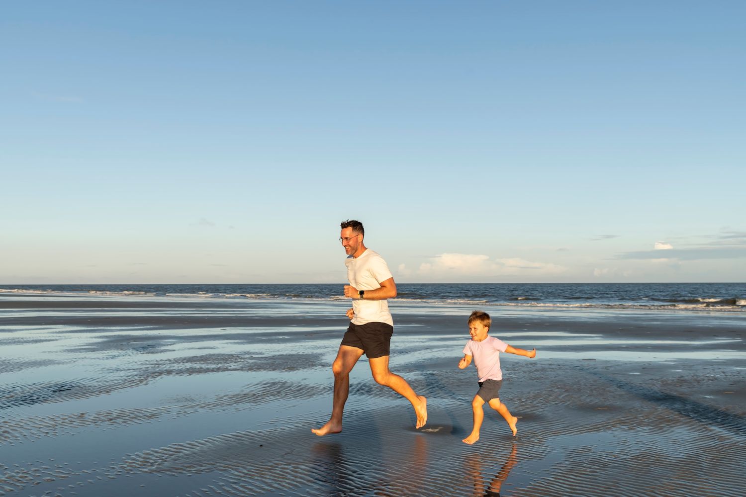 Two people jog together along a wet sandy beach at sunset with ocean waves and blue sky in the background.