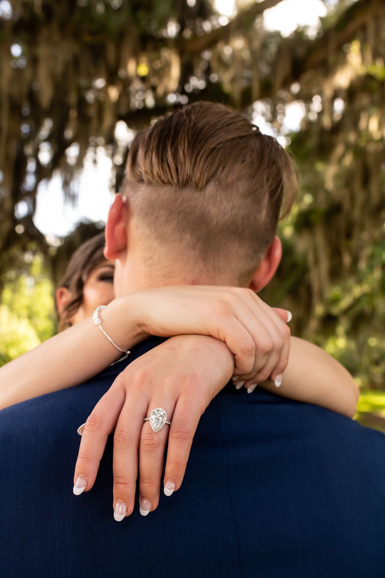 Close up of elegant engagement ring displayed on hand during romantic embrace under Spanish moss trees.