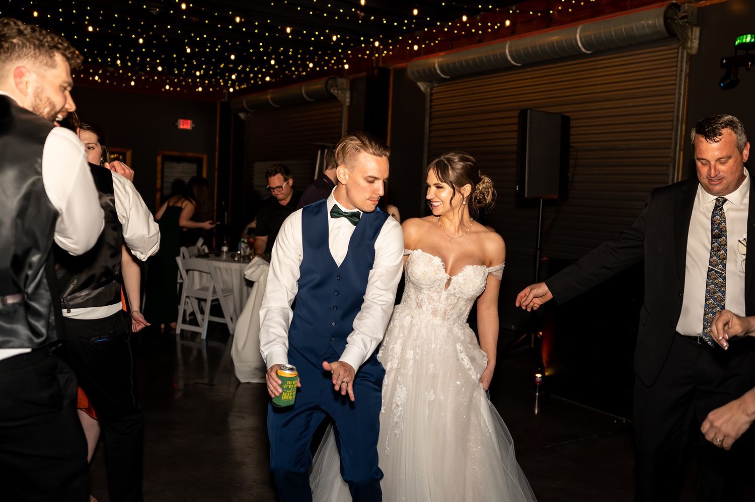 Newlyweds enjoying their first dance together at a rustic indoor reception venue under twinkling lights.