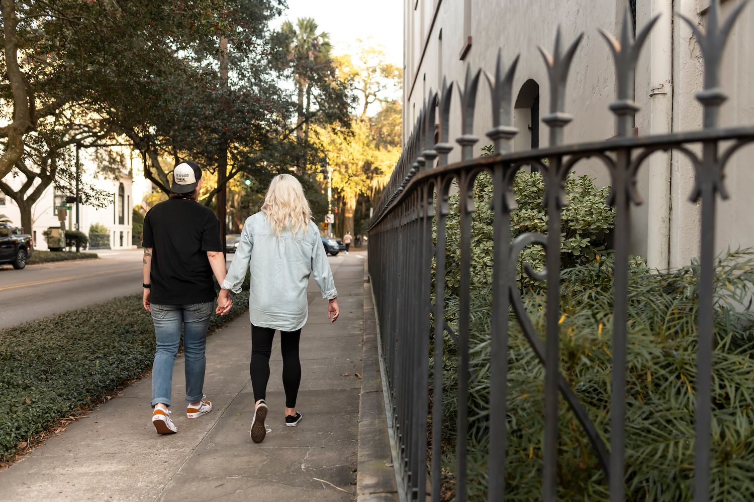 Couple walking hand in hand along a sidewalk next to decorative iron fence during sunset.