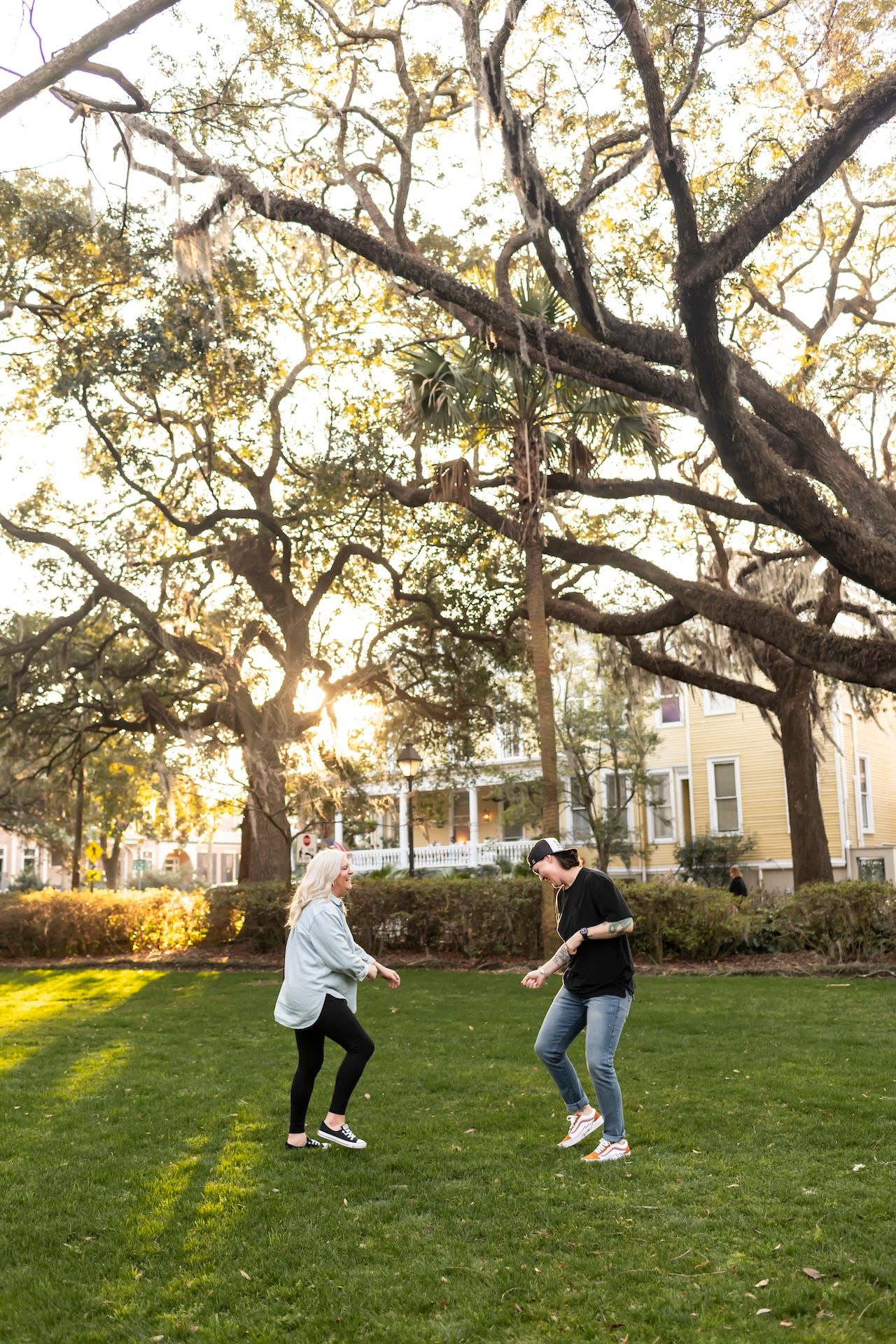 Two people playfully dance together on a grassy lawn under large oak trees at sunset.