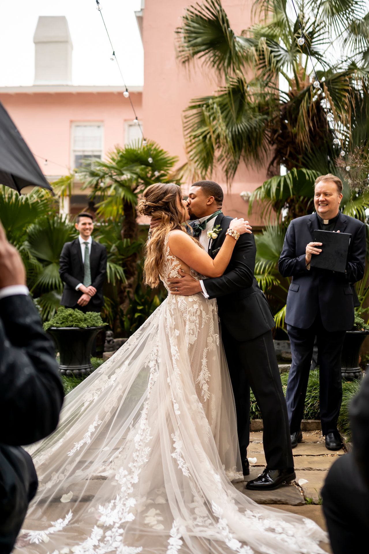 Elegant bride in flowing lace dress and groom in black suit share intimate dance moment at tropical wedding venue.