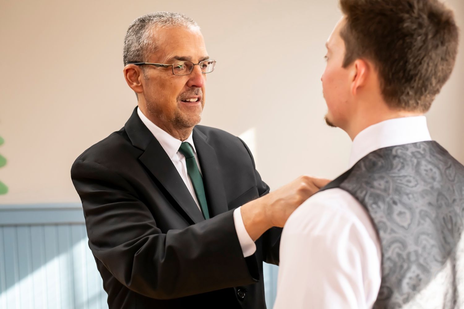 A senior professional in a dark suit adjusts the clothing of a younger person wearing a vest in an office setting.