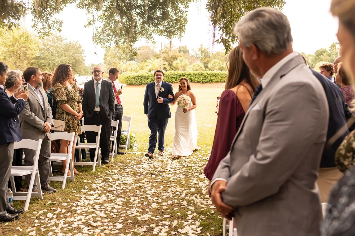 Wedding guests stand in line at an outdoor fall ceremony with scattered autumn leaves covering the ground.