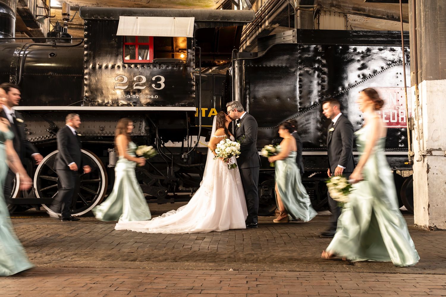 Models in flowing gowns move gracefully across a dark industrial stage during a fashion show.
