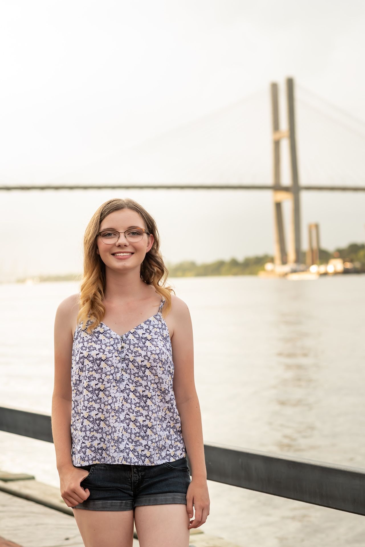 A person stands by the water smiling near a large bridge structure on a sunny day.