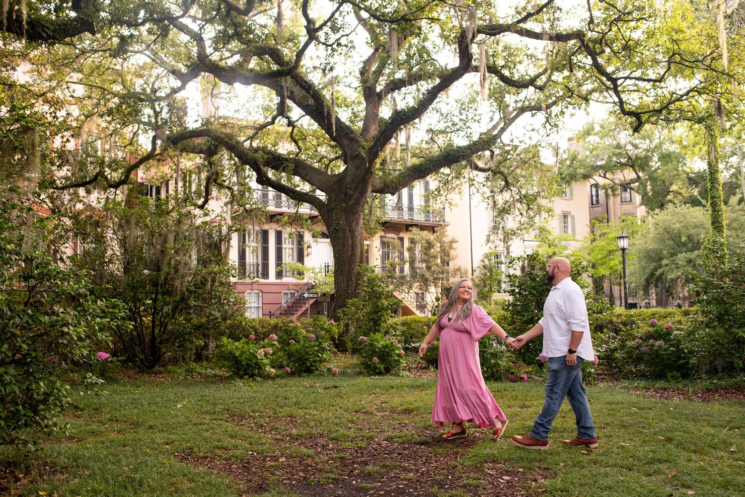 Two people walk together under a majestic oak tree in front of a historic southern home with lush gardens.