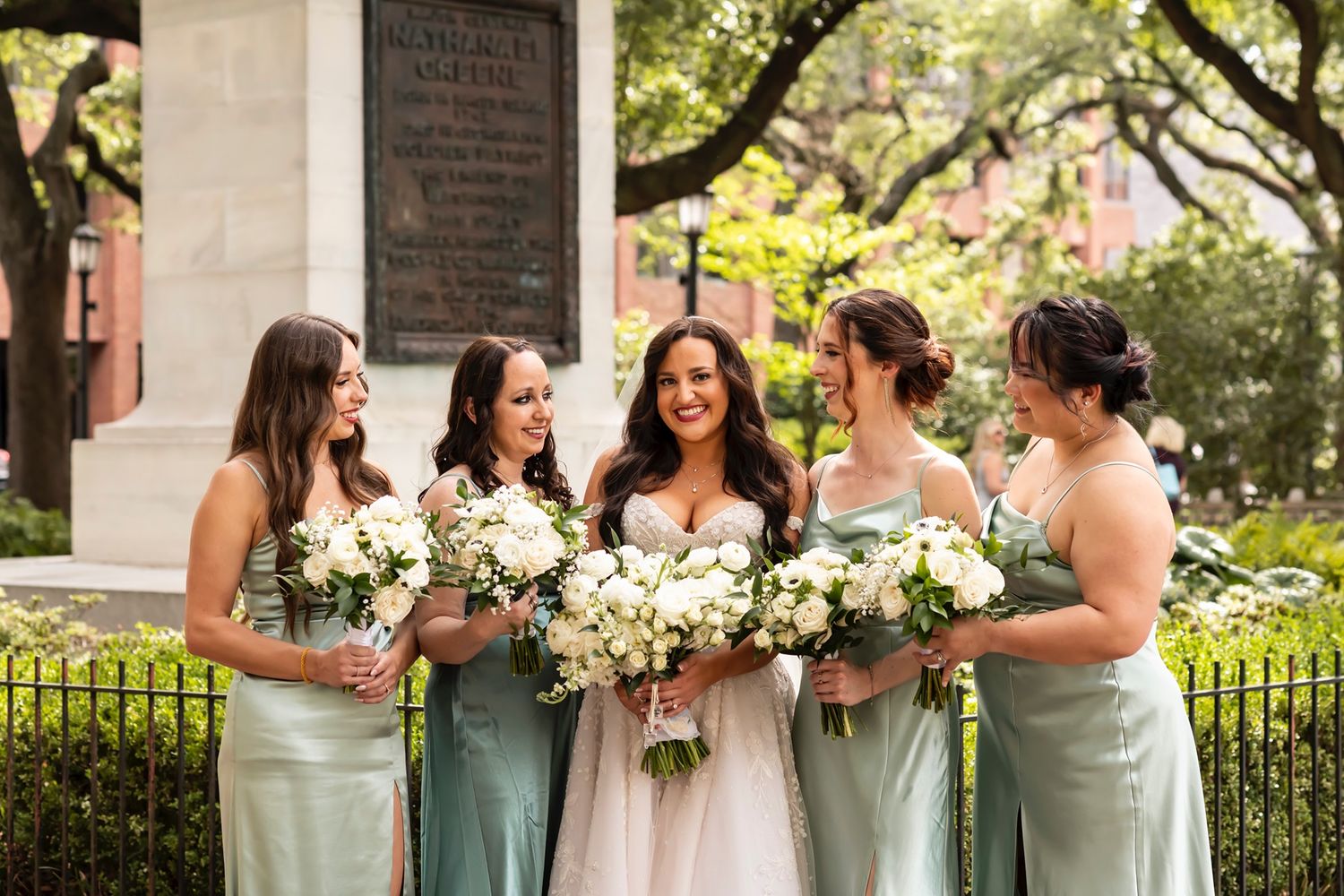 Bridesmaids in sage green dresses holding white bouquets pose together at an outdoor garden venue.