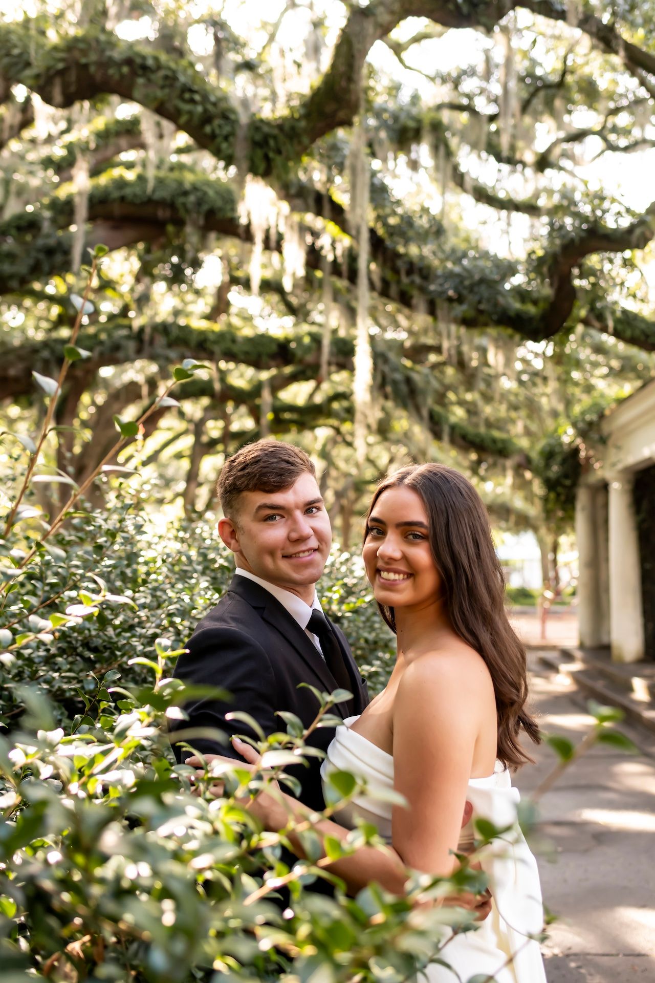 A couple poses together outdoors under Spanish moss draped oak trees in Savannah during a formal occasion.