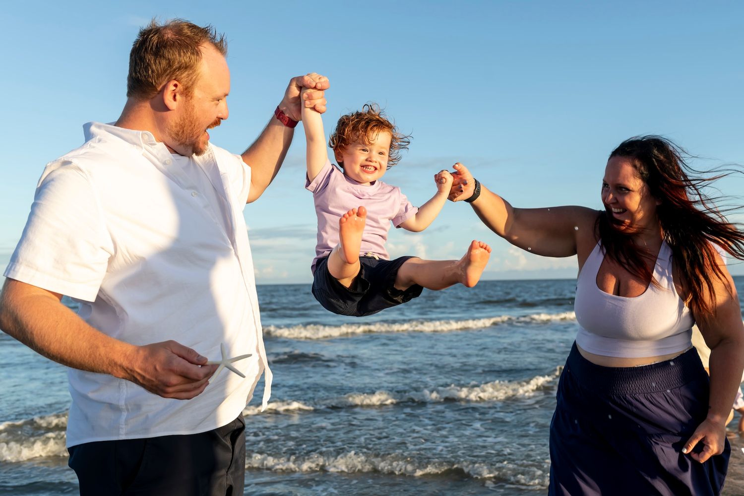 Parents holding and swinging small child together at beach with ocean waves and blue sky in background.